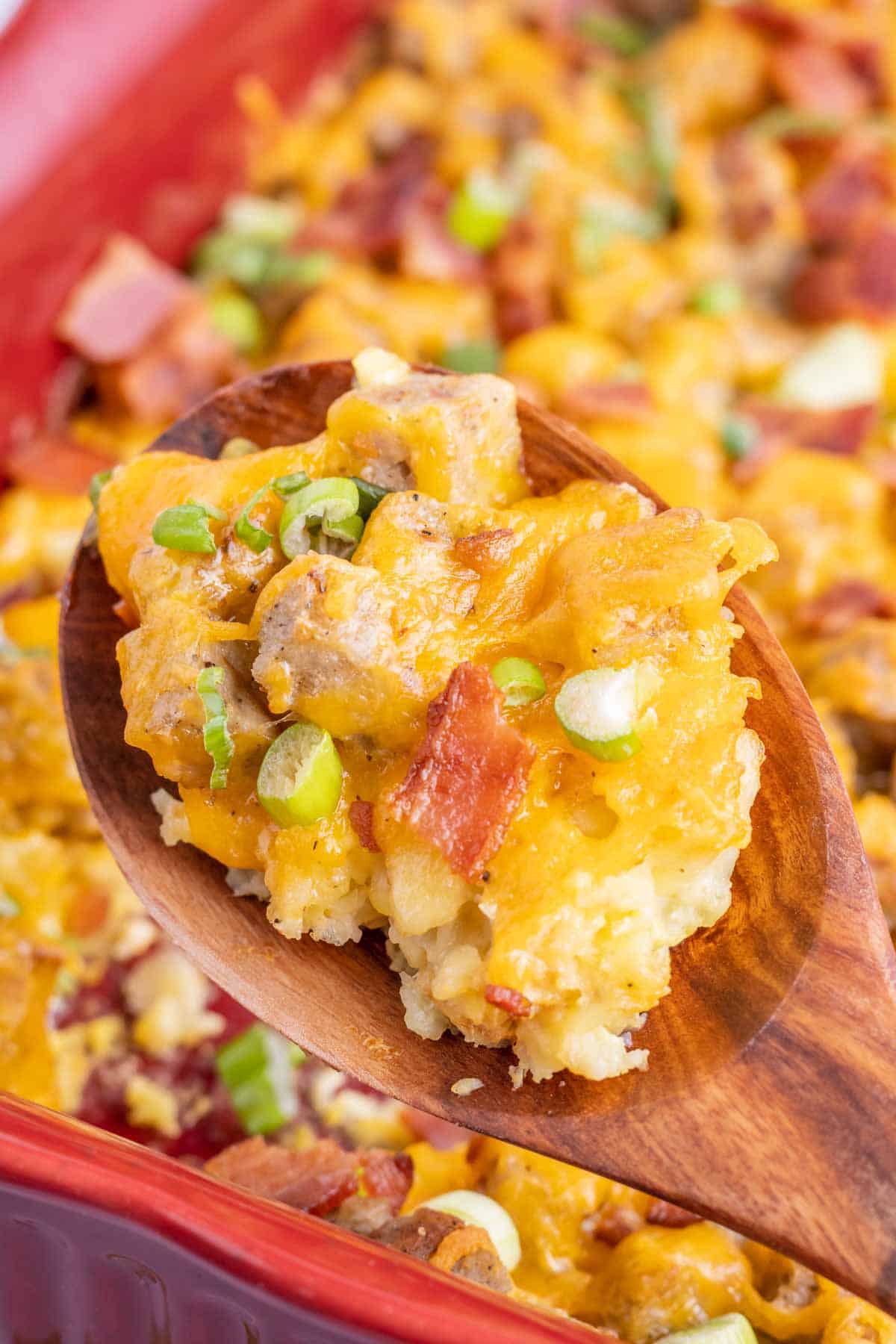 A wooden spoon holding a serving of cheesy breakfast casserole with sausage, bacon, green onions, and potatoes in a red baking dish.