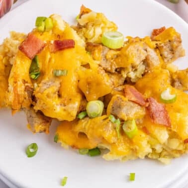 A serving of tater tot breakfast casserole with cheese, sausage, bacon, and chopped green onions on a white plate.
