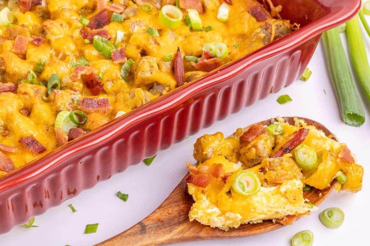 A red baking dish filled with a cheesy casserole topped with bacon and sliced green onions, with a wooden spoon holding a serving next to it.
