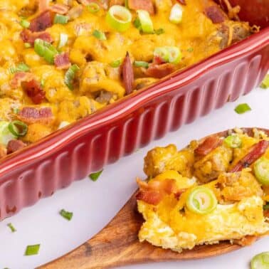 A baked tater tot casserole in a red dish topped with melted cheese, bacon, and sliced green onions, with a wooden spoon serving a portion.