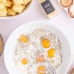 A bowl with cracked eggs, milk, pepper, and a whisk, surrounded by tater tots, garlic powder, eggshells, and ingredients on a countertop.