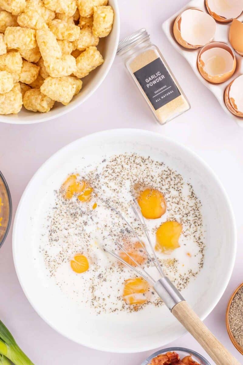 A bowl with cracked eggs, milk, pepper, and a whisk, surrounded by tater tots, garlic powder, eggshells, and ingredients on a countertop.