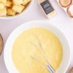 A bowl of whisked eggs with a whisk inside, surrounded by tater tots, garlic powder, cracked eggshells, green onions, and bacon on a white surface.