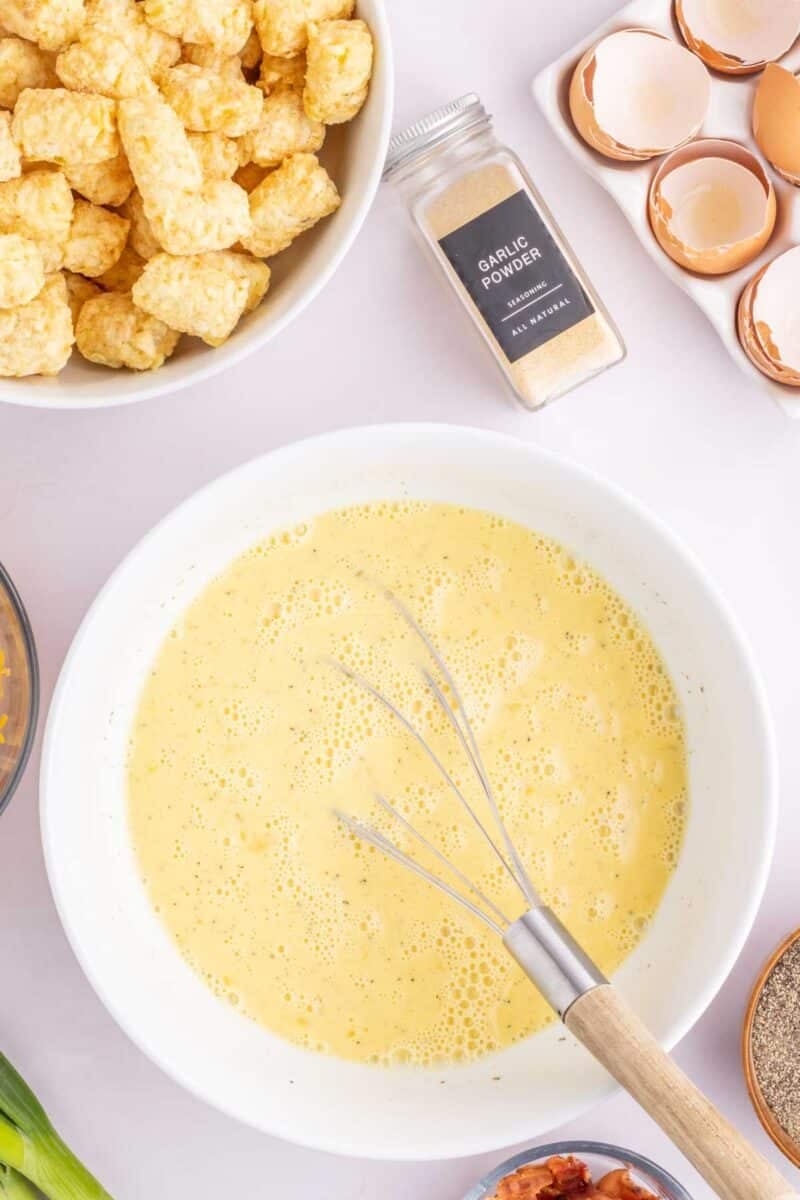 A bowl of whisked eggs with a whisk inside, surrounded by tater tots, garlic powder, cracked eggshells, green onions, and bacon on a white surface.