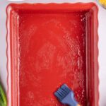 A red rectangular baking dish coated with oil, a silicone brush resting inside, surrounded by tater tots, shredded cheese, green onions, and a small bowl of liquid.