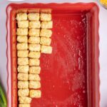 A red baking dish partially lined with rows of frozen tater tots, with other ingredients like cheese and green onions nearby.