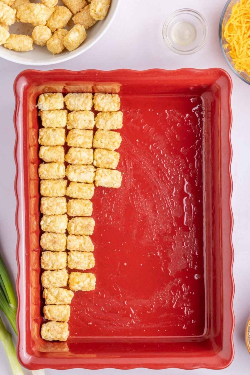 A red baking dish partially lined with rows of frozen tater tots, with other ingredients like cheese and green onions nearby.