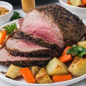 Sliced roast beef served with roasted potatoes and carrots, garnished with parsley, on a white plate.