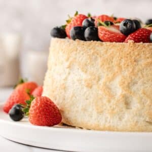 A round sponge cake topped with fresh strawberries and blueberries sits on a white plate.