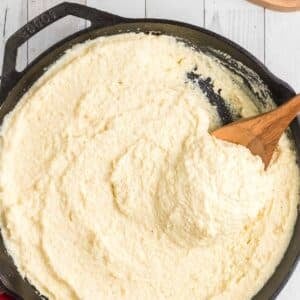 A cast iron skillet filled with creamy, baked polenta being stirred with a wooden spoon on a white wooden surface.