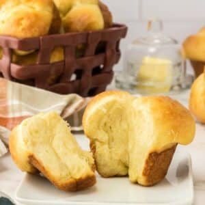 A golden brown yeast roll split open on a white plate, with a basket of more rolls and a butter dish in the background.