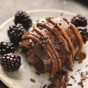 A chocolate cremeux dessert with caramel drizzle and chocolate shavings, served on a plate with fresh blackberries.