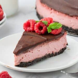A slice of raspberry cheesecake with a chocolate crust and chocolate topping, garnished with raspberries and mint, served on a white plate.
