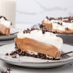A slice of chocolate cream pie with whipped topping and chocolate shavings on a plate, with more pie and a glass of milk in the background.