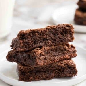Three chocolate brownies are stacked on a white plate, with a blurred glass of milk and additional brownies in the background.