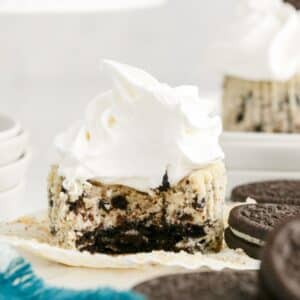 A cookies and cream cheesecake cupcake topped with whipped cream, shown with a bite taken out, next to whole sandwich cookies.