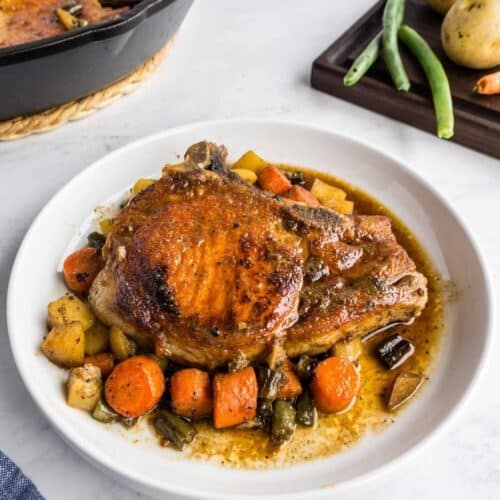 A cooked pork chop served on a white plate with carrots, potatoes, and green beans in a savory sauce.