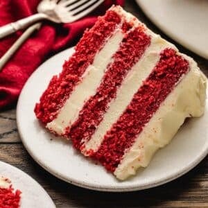 A slice of red velvet cake with three layers and white cream cheese frosting on a white plate, with a fork and red napkin in the background.