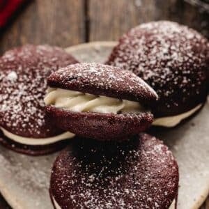 Three red velvet whoopie pies with cream filling, topped with a dusting of powdered sugar, arranged on a plate.
