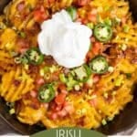 A skillet of Irish nachos topped with melted cheese, bacon, jalapeños, chopped tomatoes, green onions, and a dollop of sour cream.