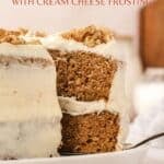 A carrot cake with cream cheese frosting and chopped nuts on top sits on a plate with a slice partially removed.