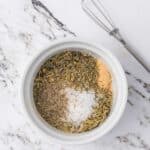 A white bowl with dried herbs, ground pepper, salt, and garlic powder on a marble surface next to a metal whisk.