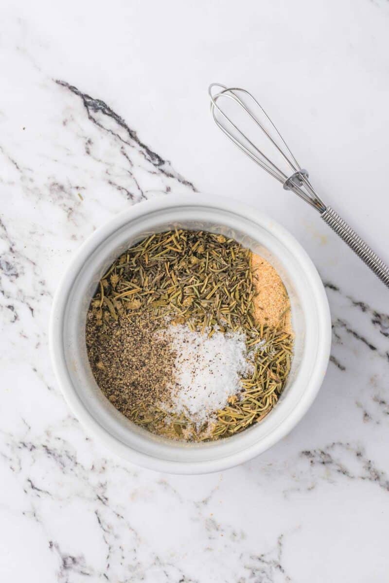 A white bowl with dried herbs, ground pepper, salt, and garlic powder on a marble surface next to a metal whisk.
