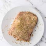 A piece of raw meat coated with dry seasoning sits on a white plate atop a marble surface.