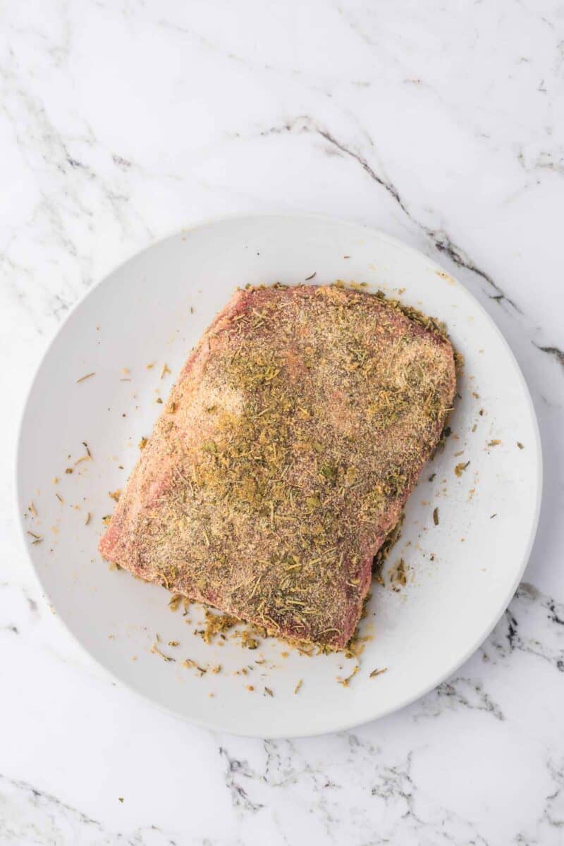 A piece of raw meat coated with dry seasoning sits on a white plate atop a marble surface.