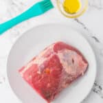 A raw beef roast sits on a white plate beside a bowl of oil and a blue basting brush on a marble countertop.