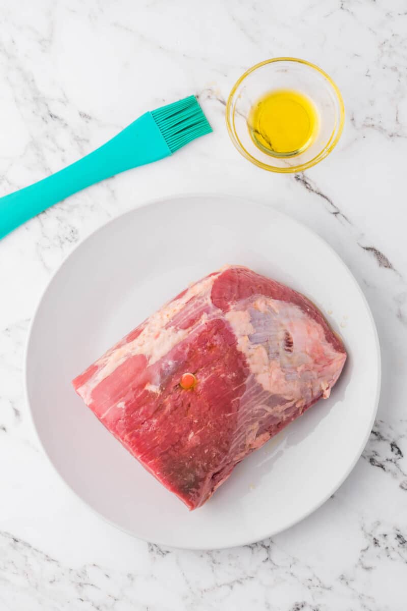 A raw beef roast sits on a white plate beside a bowl of oil and a blue basting brush on a marble countertop.