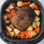 A cooked roast surrounded by chopped potatoes and carrots in an air fryer basket on a marble countertop.
