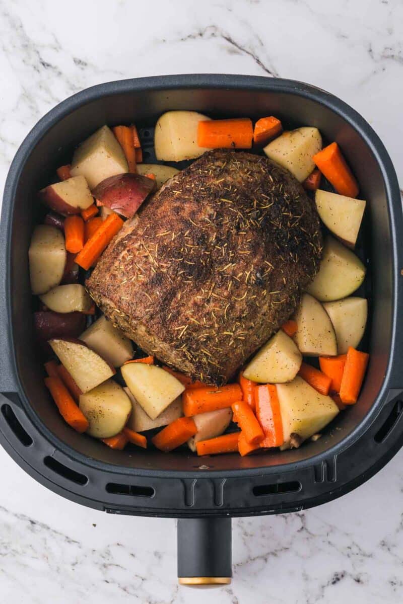 A cooked roast surrounded by chopped potatoes and carrots in an air fryer basket on a marble countertop.