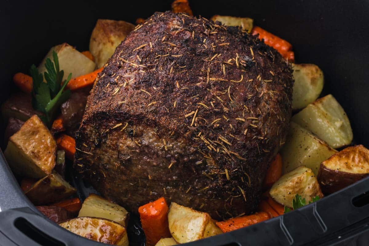 A seasoned beef roast sits in a pot surrounded by roasted potato chunks and carrot pieces.