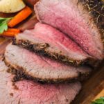 Sliced roast beef with a pink center arranged on a wooden cutting board, served with roasted potatoes and carrots.