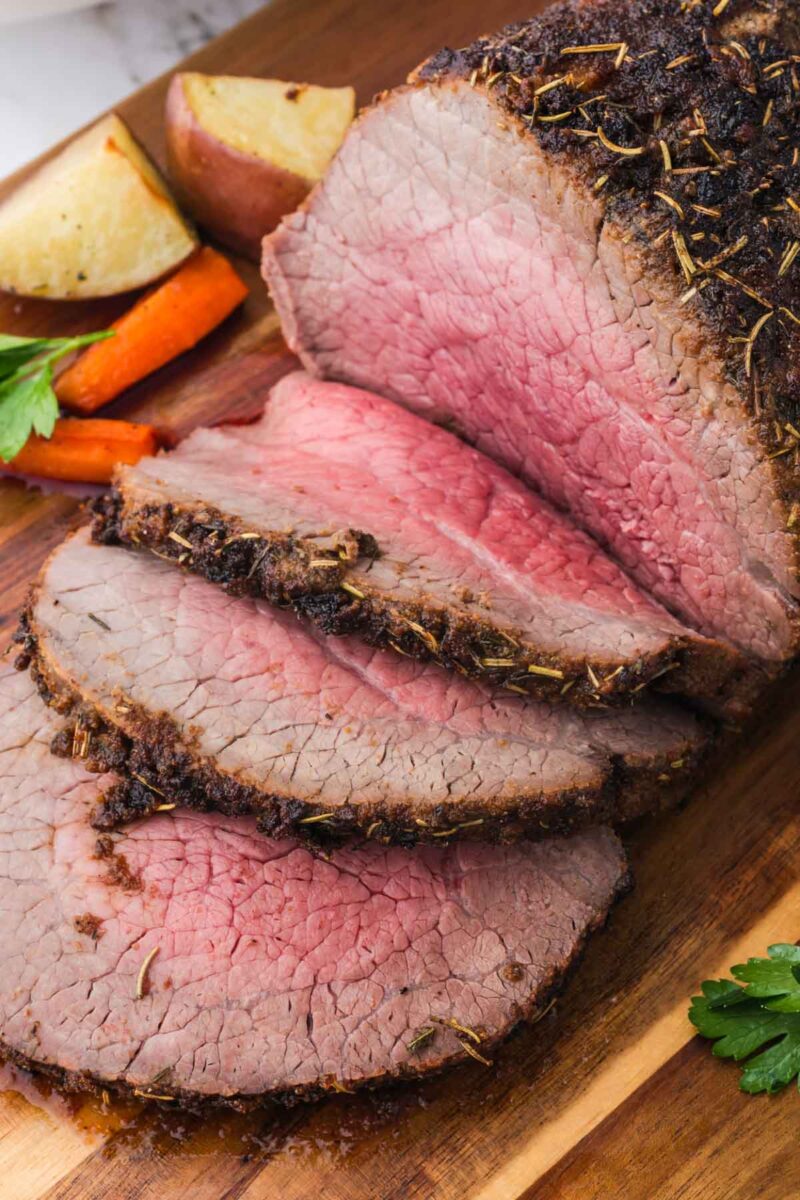 Sliced roast beef with a pink center arranged on a wooden cutting board, served with roasted potatoes and carrots.