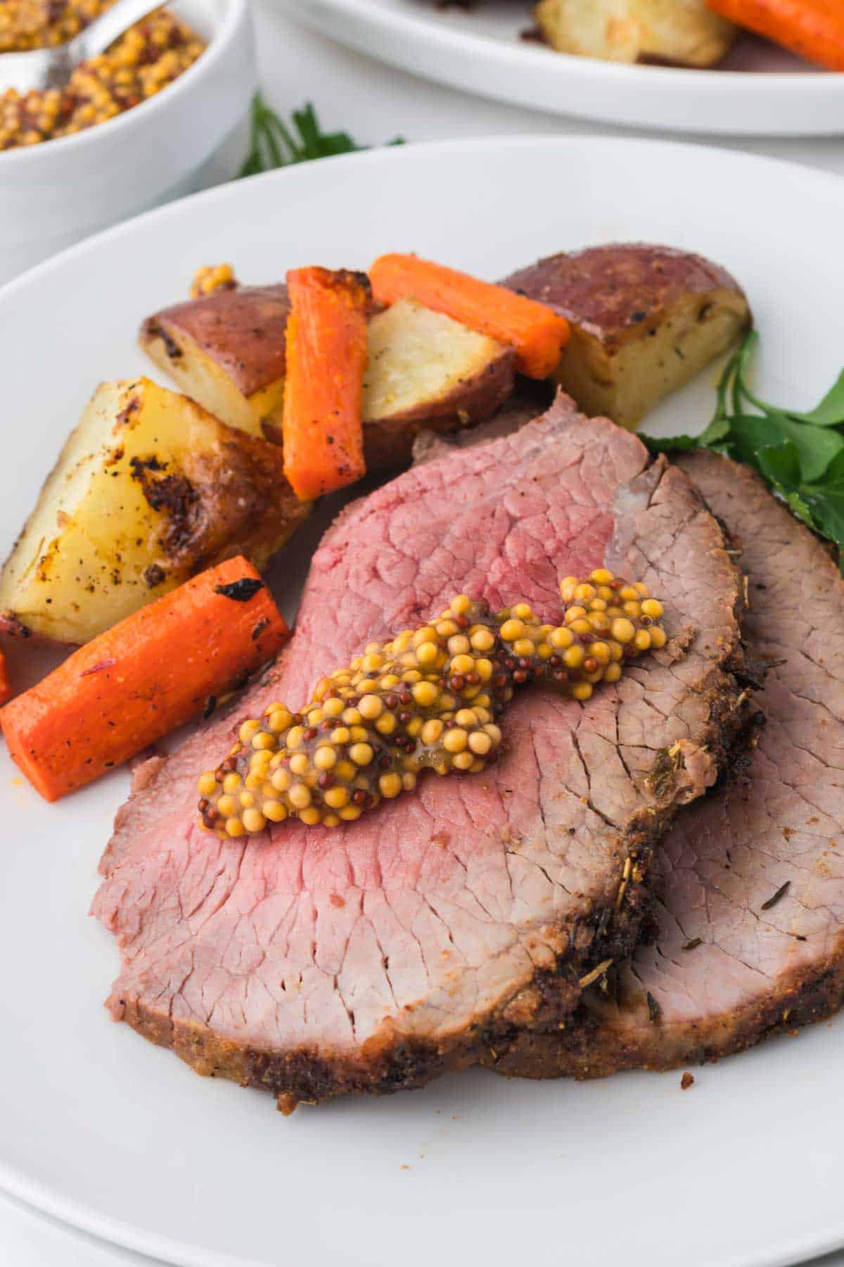 Two slices of roast beef topped with whole grain mustard, served with roasted potatoes and carrots on a white plate.