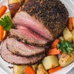 Sliced roast beef served on a white plate with roasted potatoes, carrots, and parsley garnish.