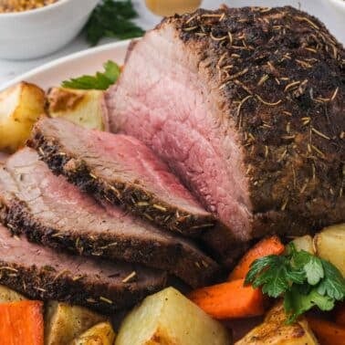 Sliced roast beef with a seasoned crust, served with roasted carrots and potatoes on a white plate.