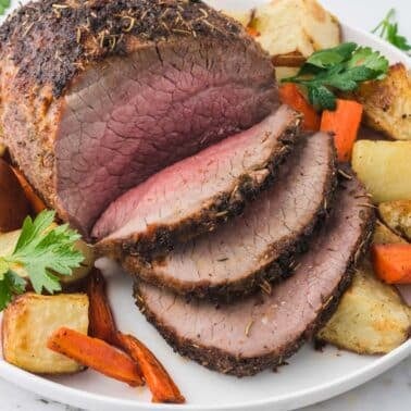 Sliced roast beef on a white plate, served with roasted potatoes, carrots, and garnished with fresh parsley.