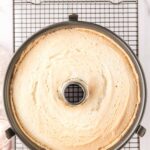 A freshly baked angel food cake in a tube pan sits on a cooling rack, viewed from above.