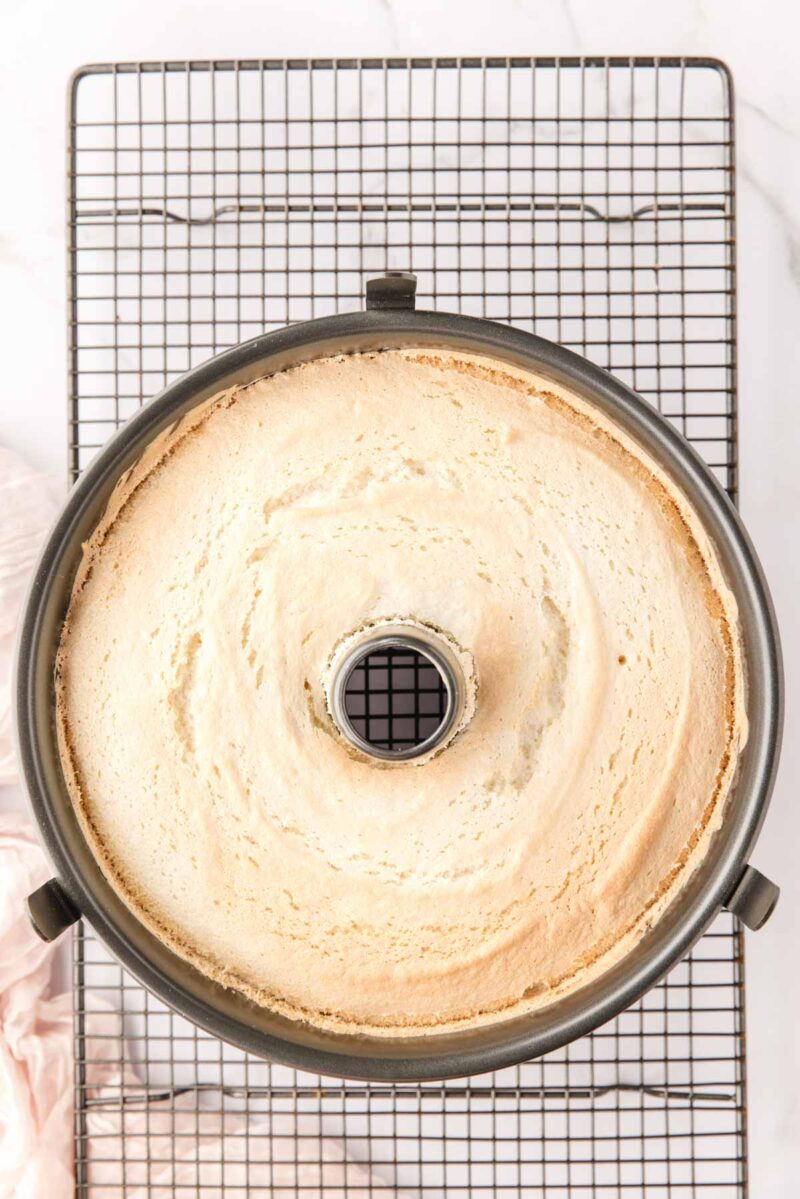 A freshly baked angel food cake in a tube pan sits on a cooling rack, viewed from above.