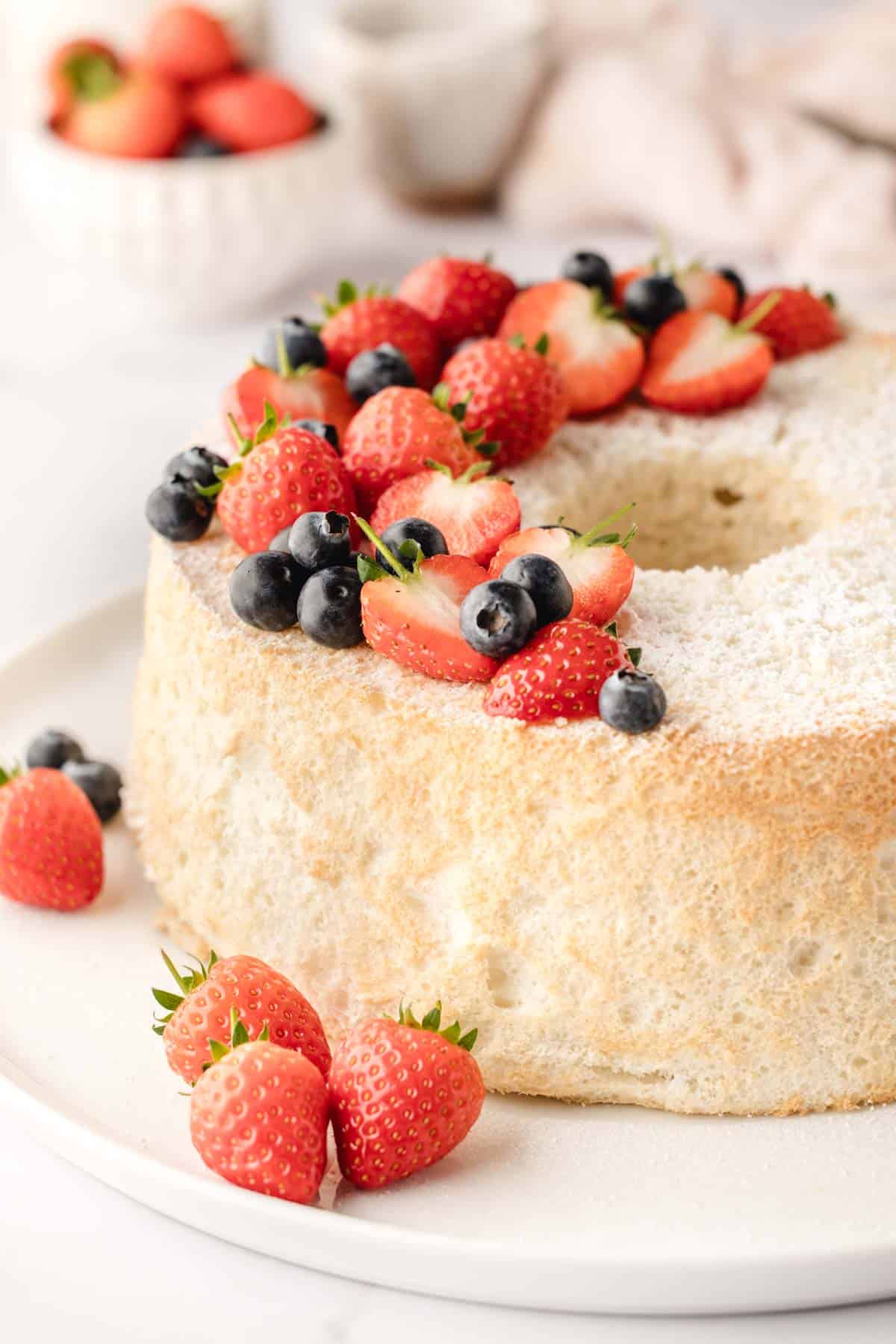 A round sponge cake dusted with powdered sugar, topped with fresh strawberries and blueberries, sits on a white plate.
