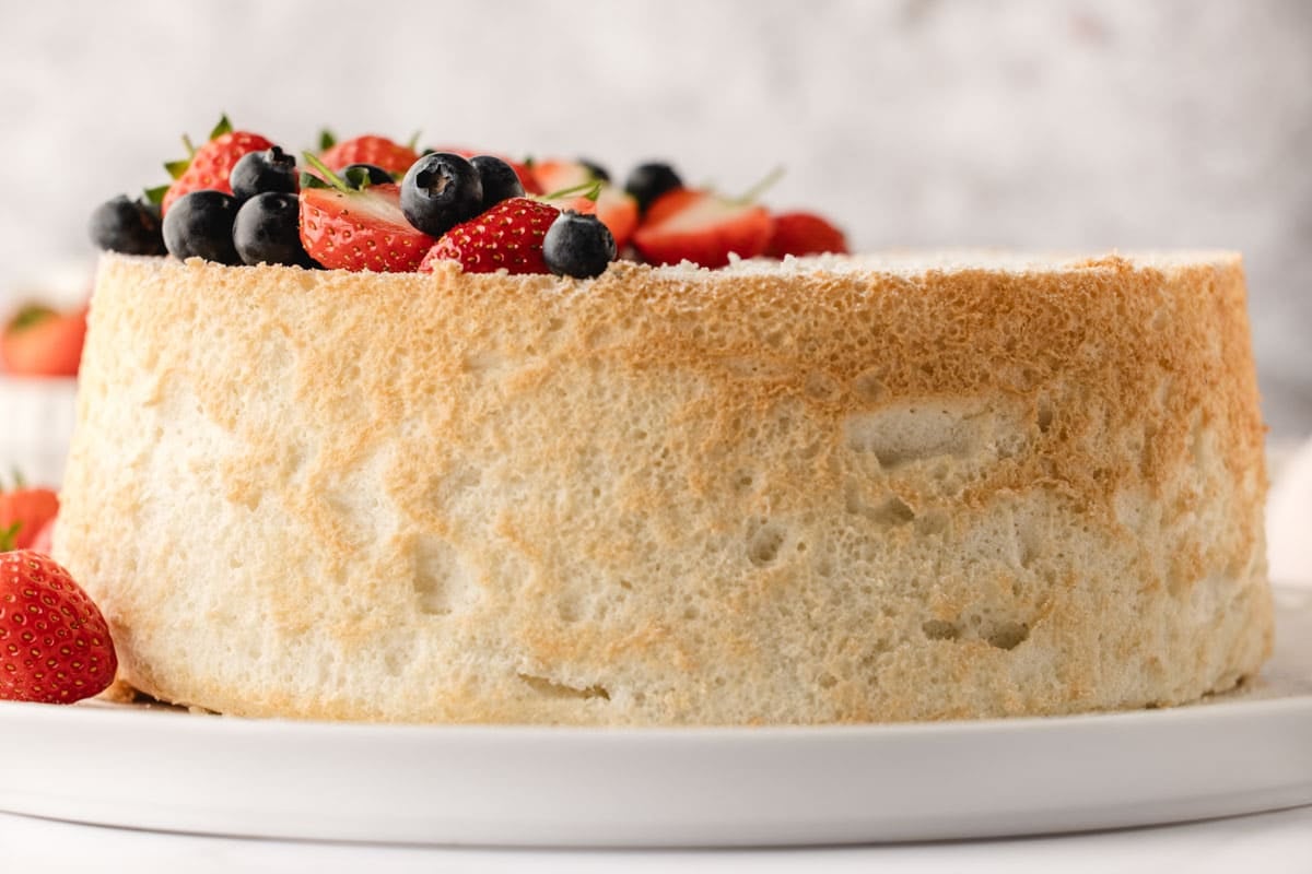 A plain angel food cake on a white plate, topped with fresh strawberries and blueberries.