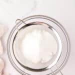 A metal sieve containing white powdered sugar rests over a glass bowl on a white marble surface.
