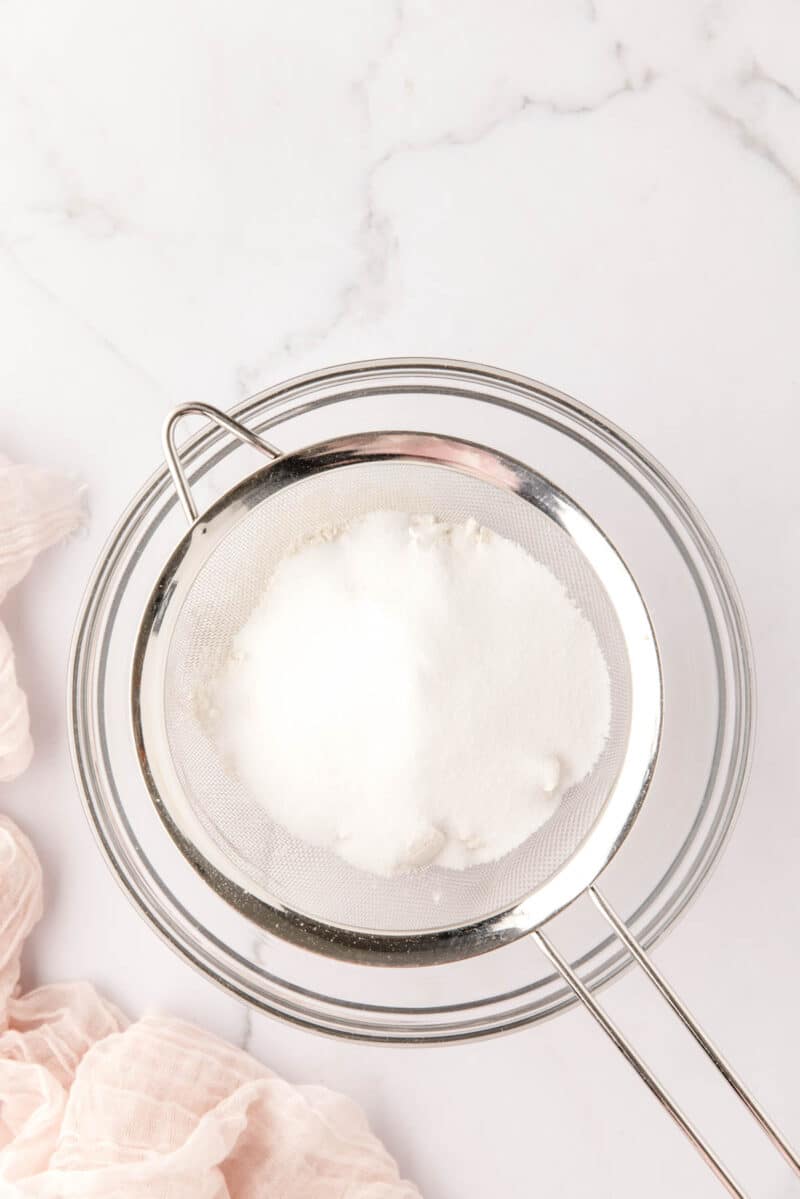 A metal sieve containing white powdered sugar rests over a glass bowl on a white marble surface.