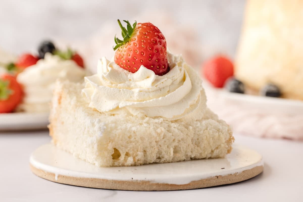 A slice of white sponge cake topped with swirled whipped cream and a fresh strawberry, served on a small round plate.