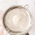 A metal sifter with flour inside is placed over a silver mixing bowl on a white marble surface.
