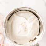 A metal mixing bowl filled with smooth, whipped cream on a white marble surface, with a pale pink cloth nearby.