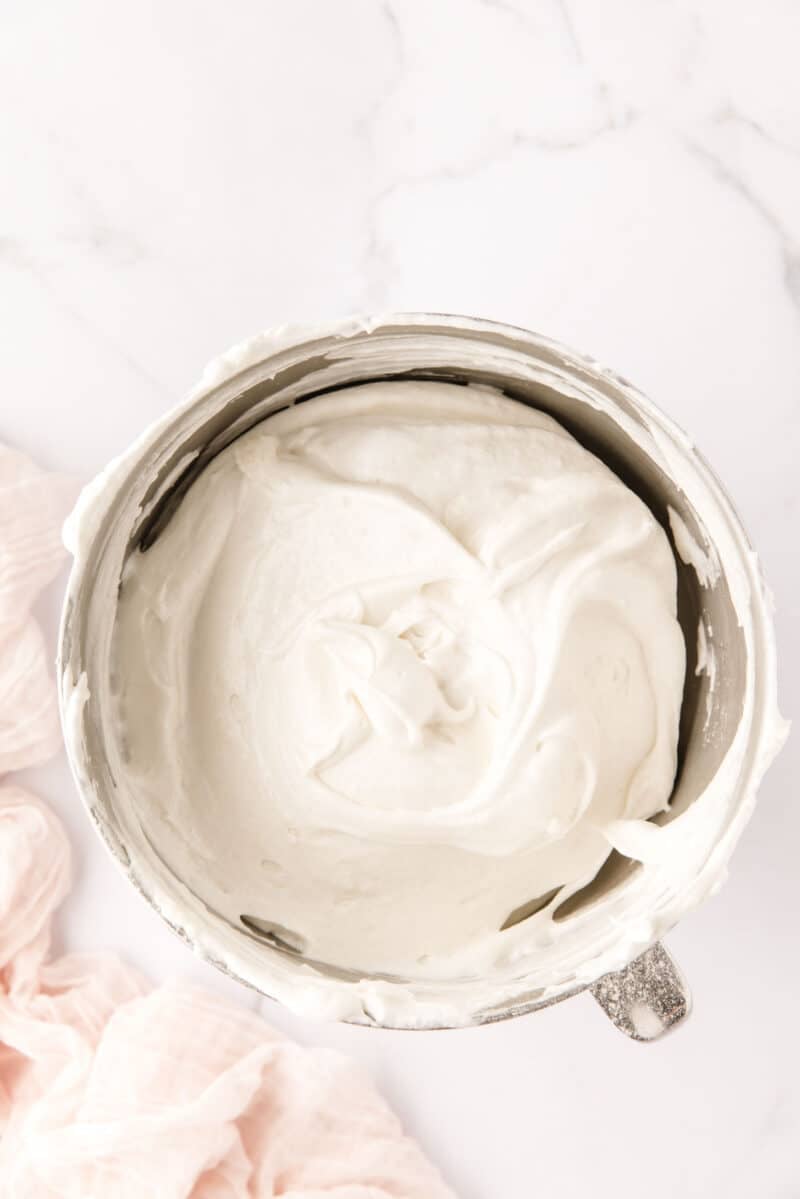 A metal mixing bowl filled with smooth, whipped cream on a white marble surface, with a pale pink cloth nearby.